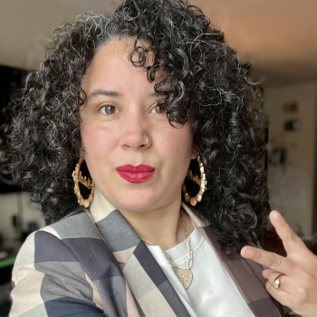 Photograph of a person with curly dark brown hair, hoop earrings, red lipstick, a brown blazer and showing fingers as a peace sign. 