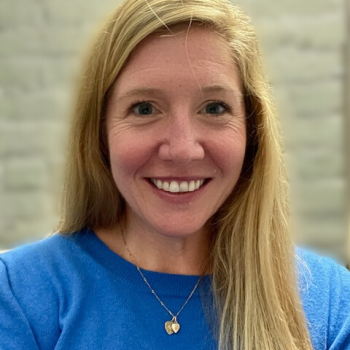 A white woman with long blonde hair and blue eyes, wearing a bright blue sweater and thin gold necklace, facing the camera and smiling in front of a gray background.. 