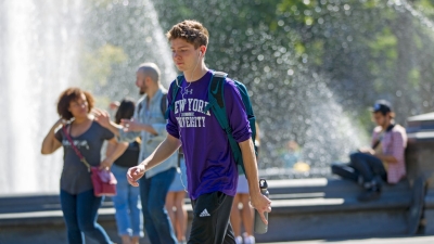 An undergrad student wearing an NYU T shirt walks past Washington square fountain
