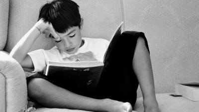 A child sitting on a couch and reading a book with his hand on his forehead