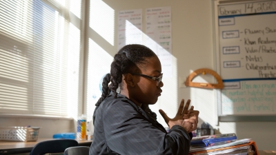 A Black girls sits alone in a classroom staring down at her assignment