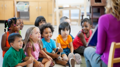 A photo of nine children sitting on the floor of a classroom and watching an adult present a book. The adult has their back to the camera and the text of the book is not visible.