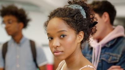 A standing girl student stares at the camera in a classroom with a background of other standing students