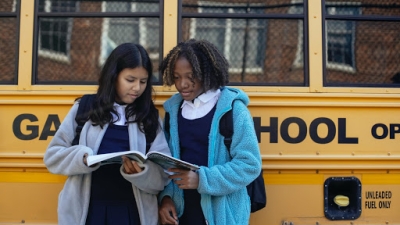 Two girls hold a textbook and look at the pages as they stand in front of a school bus