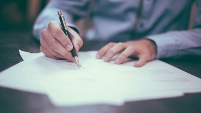 A photo of a man in a blue button down shirt using a pen to write on paper. The writing on the paper is out of focus, and the man's face is obscured.