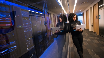 A photo of a woman with a laptop leaning against a glass wall that separates her from a collection of computer servers