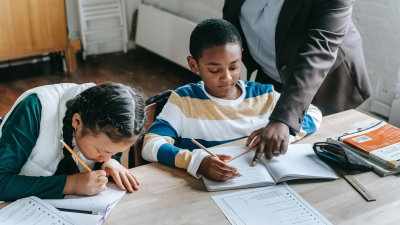 A photo of a teacher helping two children, sitting at a desk, with their school work