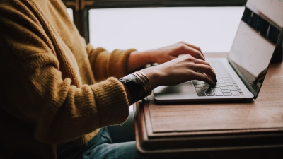 A stock image of a person in a sweater typing on a laptop. Their face is cropped out of the photo.