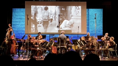 The NYU Contemporary Music ensemble featuring strings, woodwinds, brass, and percussion perform live music synchronized with a film projected on a large screen