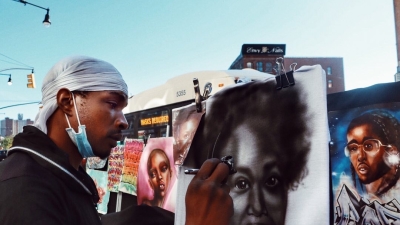 Street artist airbrushing a portrait of a Black woman.