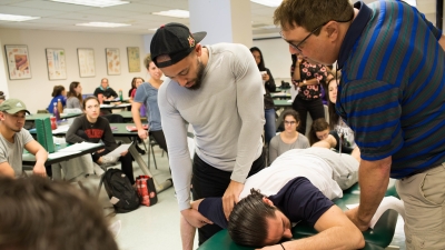 A man, laying facedown on a padded medical table, has his arm manipulated by a physical therapy student while an instructor and classmates look on.