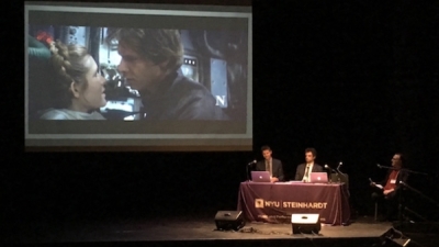 Screen showing Star Wars clip with seminar speakers sitting at desk in front of it discussing