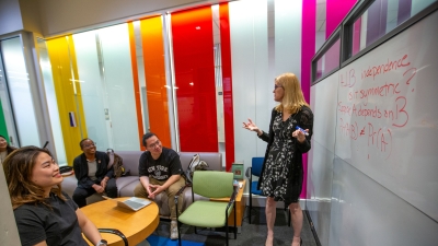 Professor Jennifer Hill by white board, explaining an equation to 3 sitting students 