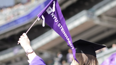 NYU graduate in cap and gown waves a Steinhardt pennant