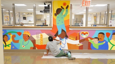 Man paints a colorful mural in a hospital. The mural depicts healthcare workers and patients.