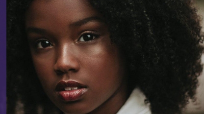 Image captures a Black girl with curly black hair looks pensively into the camera.
