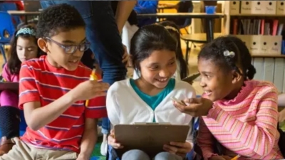 Image captures 3 elementary school students seated together in a classroom. The students are in the midst of working together to complete a classroom ag