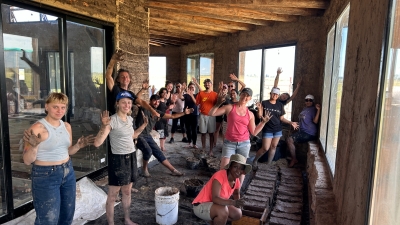 A group of students smiles and shows their muddy hands; they stand inside a partially-built mud structure