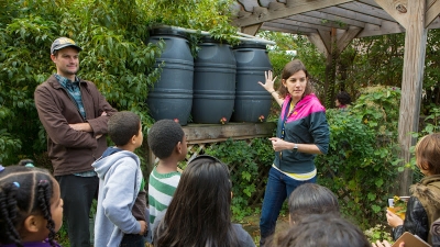 Two ECE students talking to children in front of a set of barrels.