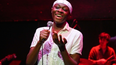 Student singing into microphone with drummer in the background