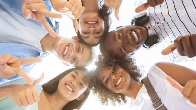 A multicultural group of five teenagers standing in a circle, viewed from a low-angle perspective with a bright sky in the background. They are smiling and gesturing with thumbs up and pointing fingers. One teen is wearing headphones around their neck, and another has curly hair. The group displays diverse hairstyles, skin tones, and clothing styles, reflecting their varied cultural backgrounds.