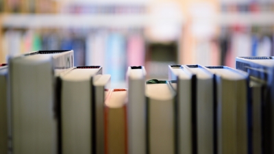 The image captures a view from inside a library shelf, looking outward through the gap between upright books. The focus is on the tops and spines of the books in the foreground, while the rest of the library, including shelves and potentially other books, is blurred in the background.