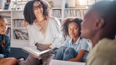 The image shows a woman with curly hair and glasses wearing a white blouse with intricate embroidery at the neckline. She is sitting and holding an open book. Around her are three children: one with braided hair, one with curly hair, and one with short hair. They are seated on the floor in a library. Behind them are shelves filled with books, and sunlight is coming through a window in the background.