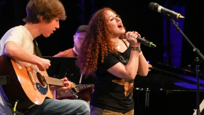 Student singing on mic while another student accompanies her on guitar