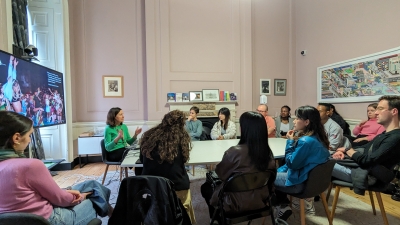 Students gathered around a classroom table and listening to an instructor speak