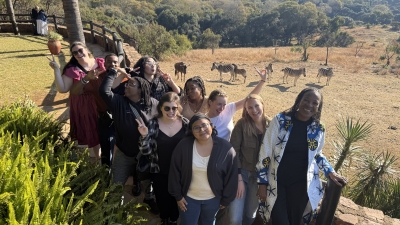A group of students and their faculty taking a photo nearby a heard of Zebras in the background.