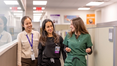 Three students, two in scrubs, walking and chatting in a hallway.