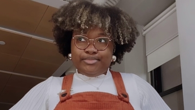Photo of Imani posing with her hands on her hips, wearing a white long sleeve top and orange overall dress