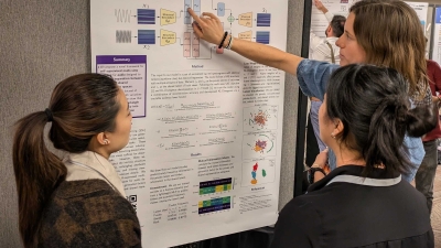 Two members of MARL point to their poster presentation, explaining the findings to a conference attendee. The project they are presenting is "Self-Supervised Mult-View Learning for Disentangled Music Audio Representations"