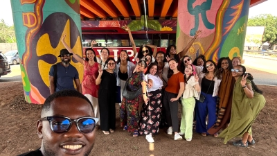 A smiling man in sunglasses takes a selfie while a group of students poses in the background under a brightly-painted bridge
