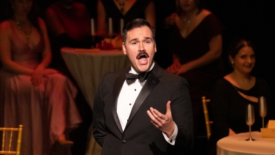 Student with mustache wearing a tuxedo singing opera on the stage with full cafe tables behind him