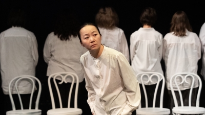 A student bending over a little; behind her are four white chairs and actors with their backs to her