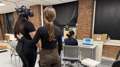 Participant stands on balance board while wearing VR headset, while one research assistant supports them with a hand on their back, and another research assistant operates a computer nearby