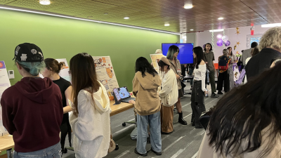 Students navigate around tables with boards and computers showcasing student work. 