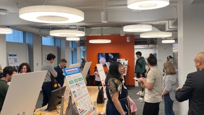 Students stand before research posters at tables in a bustling room. 