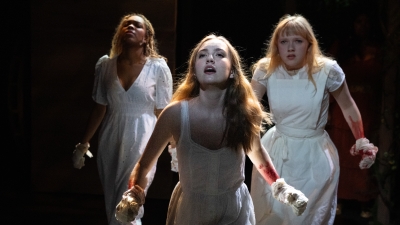 Three female students in white dresses on stage singing a dramatic song