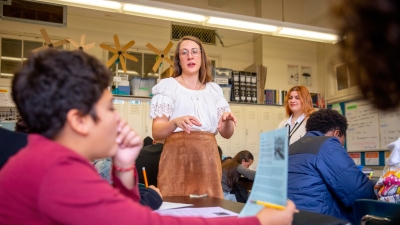 A teacher in the center of the classroom addresses a student holding a handout. 