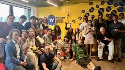 A group of students sits in front of a yellow wall that reads "Warner Music Group"