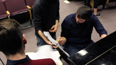 Three musicians standing at a piano working on a piece of music together.