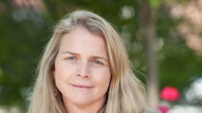 Headshot of Meredith Rowe wearing a short-sleeved red top and smiling slightly, with a softly blurred outdoor background.