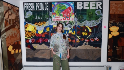 Charis stands in a gallery with one hand in their pants pocket, in front of a large scale multimedia artwork