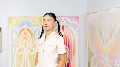Shuling wears a cream colored jumpsuit and blue boots. Her hair is styled into two pigtails and she stands in a brightly lit room in front of her ethereal paintings.