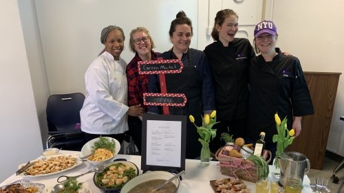 A group of students stand in front of a table of foods they prepared as a team.