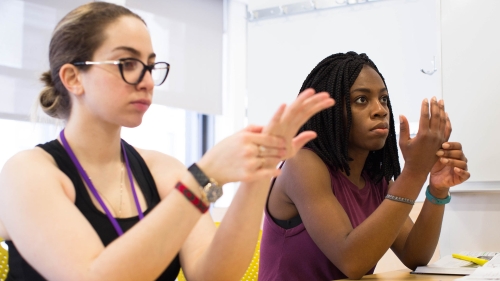 Two students holding one of their hands while seated at a table in a classroom.