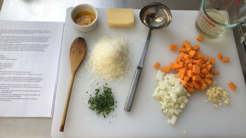 A cutting board with the elements of a recipe on top: shopped onions, carrot, garlic, herbs, grated cheese, oil, and spoons.