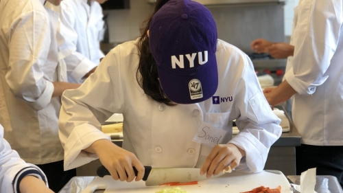 A student wearing a purple NYU hat chopping vegetables on a cutting board.